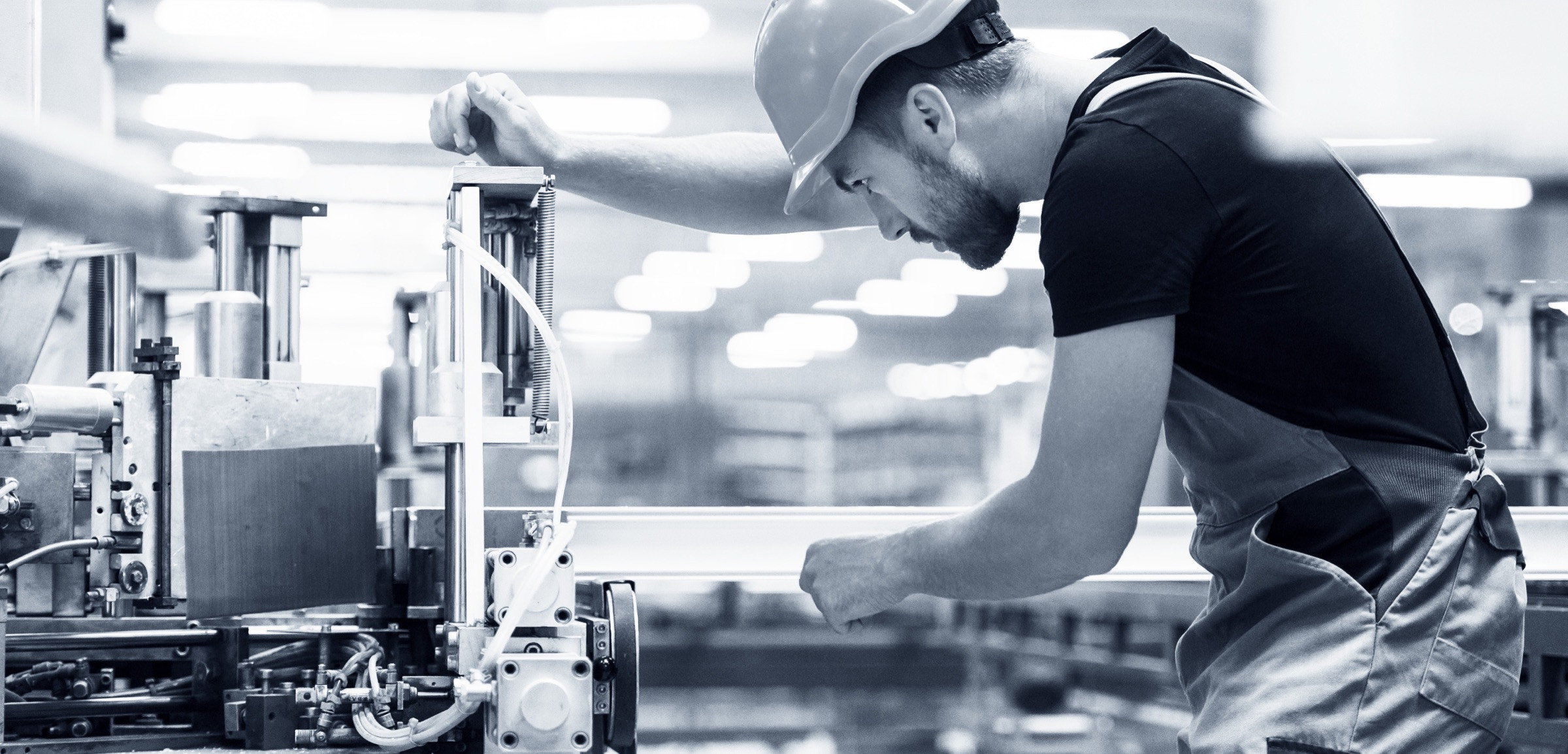 A worker in a hard hat and overalls leaning over machinery on a factory floor